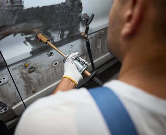 Man in special suit sitting near car, holding special tool and removing paint from the side door