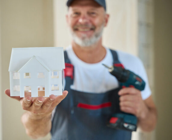 Photo of smiling construction worker in baseball cap and overalls holding house model in one hand and drill in other