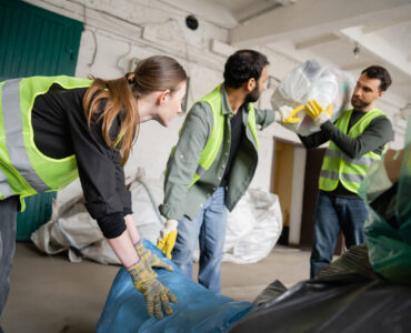 Young female worker in protective vest and gloves holding plastic bag near blurred multiethnic colleagues in waste disposal station, garbage sorting and recycling concept