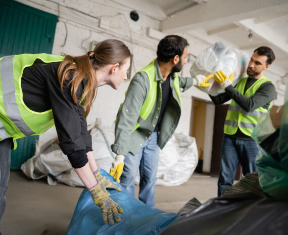Young female worker in protective vest and gloves holding plastic bag near blurred multiethnic colleagues in waste disposal station, garbage sorting and recycling concept